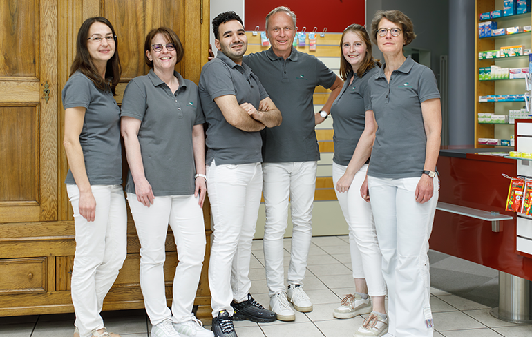 Das Team der Trierweiler Apotheke in Trierweiler steht in der Apotheke von den Medikamentenregalen.