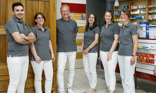 Das Team der Trierweiler Apotheke in Trierweiler steht in der Apotheke von den Medikamentenregalen.